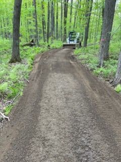 Excavator grading a new dirt trail through a lush green forest in spring.