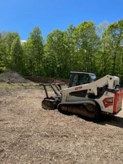 Bobcat skid steer loader on a cleared patch of land surrounded by greenery.