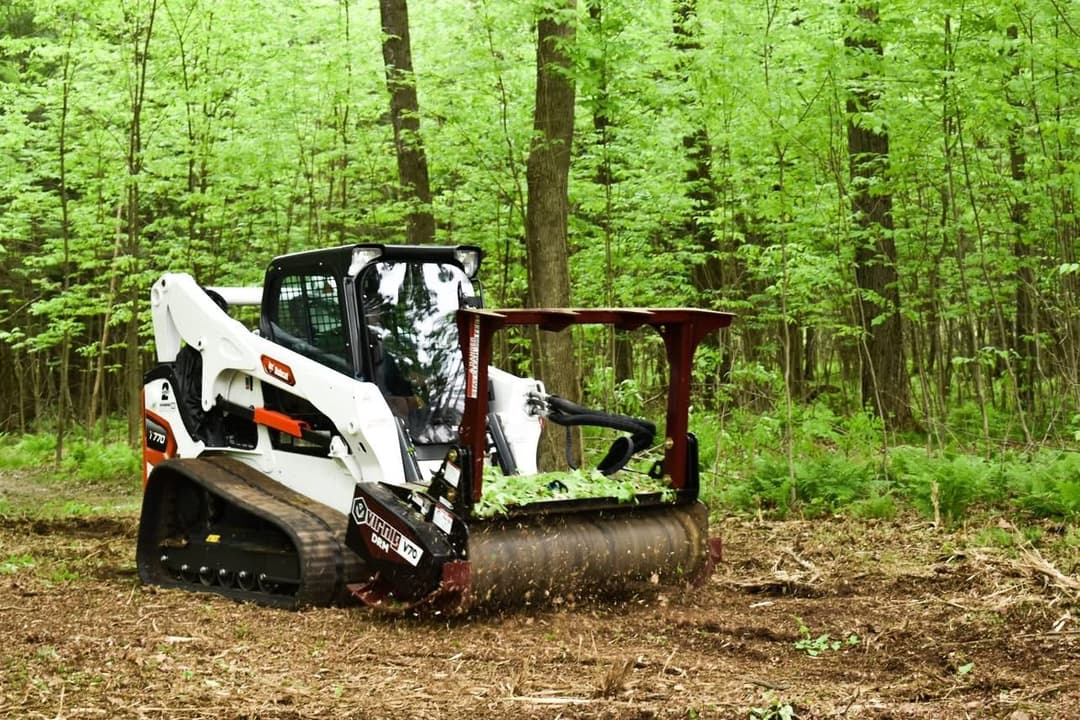 Skid steer with brush mower clearing underbrush in a lush green forest.