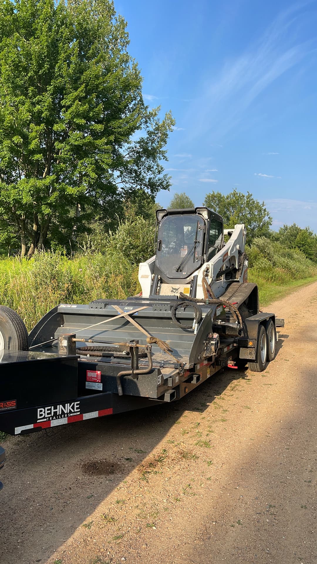 Bobcat skid steer loader on a flatbed trailer parked on a dirt road with trees in the background.