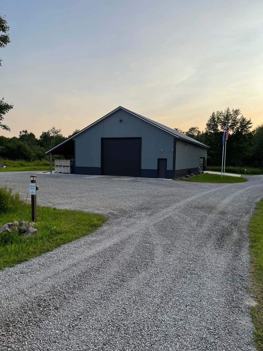 Gray metal barn with a gravel driveway surrounded by greenery at sunset.