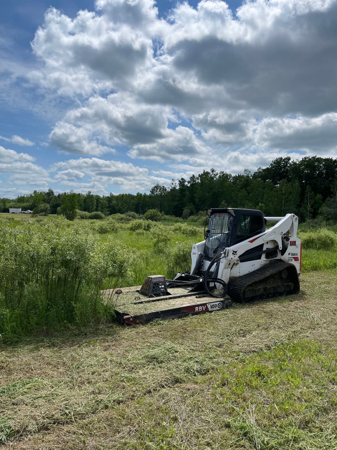 Bobcat skid steer clearing grass in a lush green field under a cloudy sky.