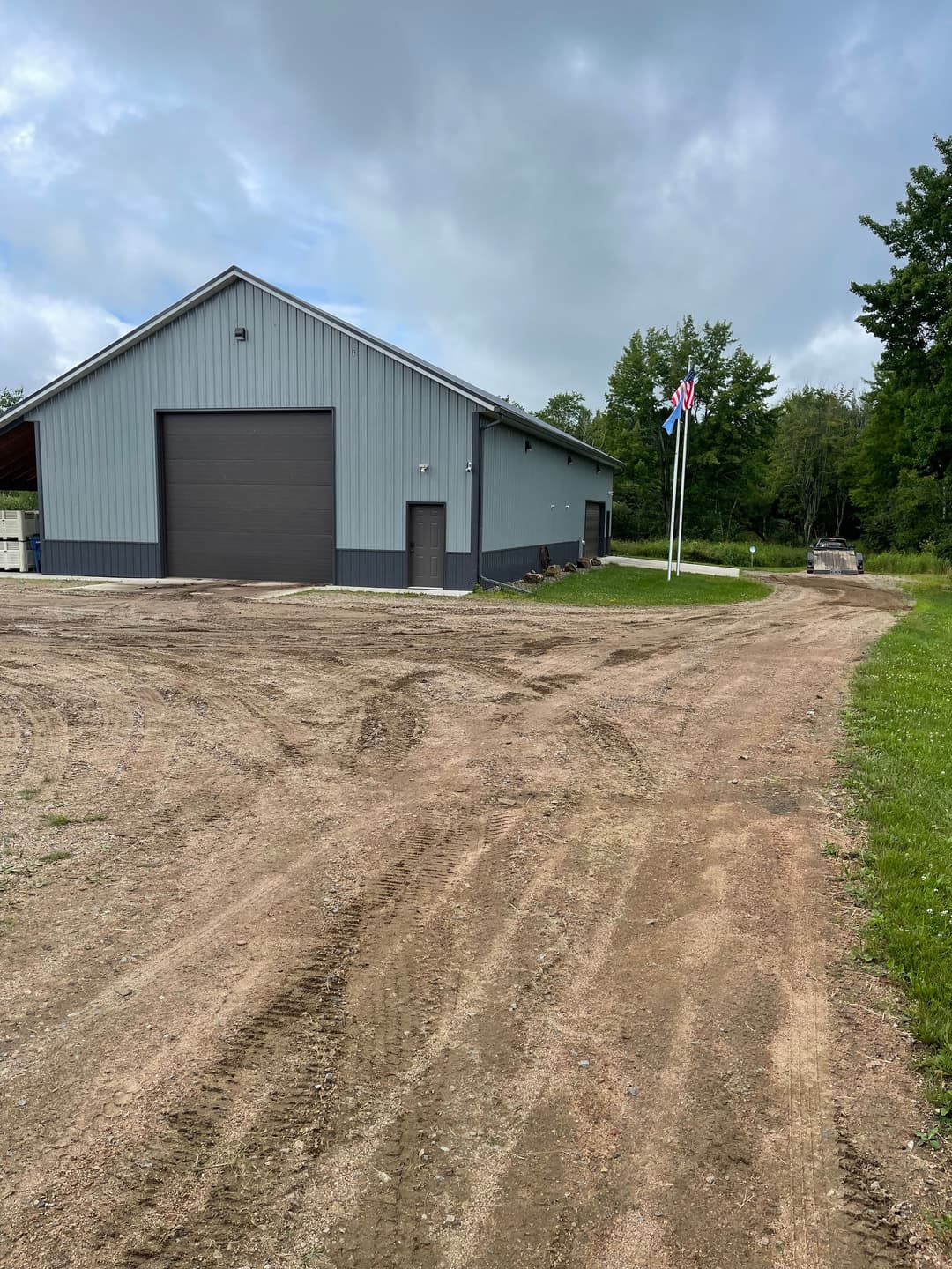 Gray metal building with a large garage door, flags, and a gravel driveway in a rural setting.