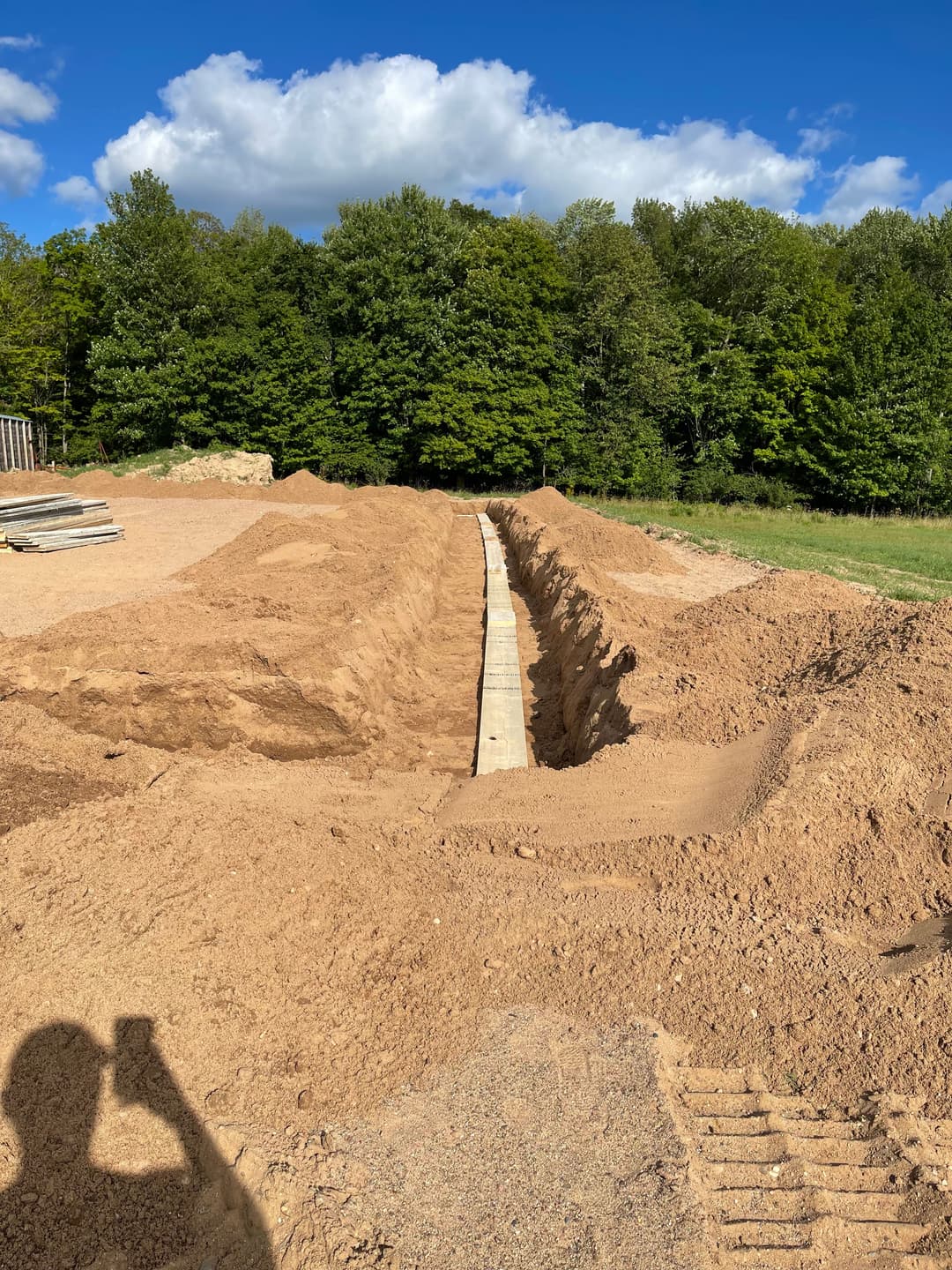 Excavated trench with wooden planks, surrounded by sandy soil and lush green trees.