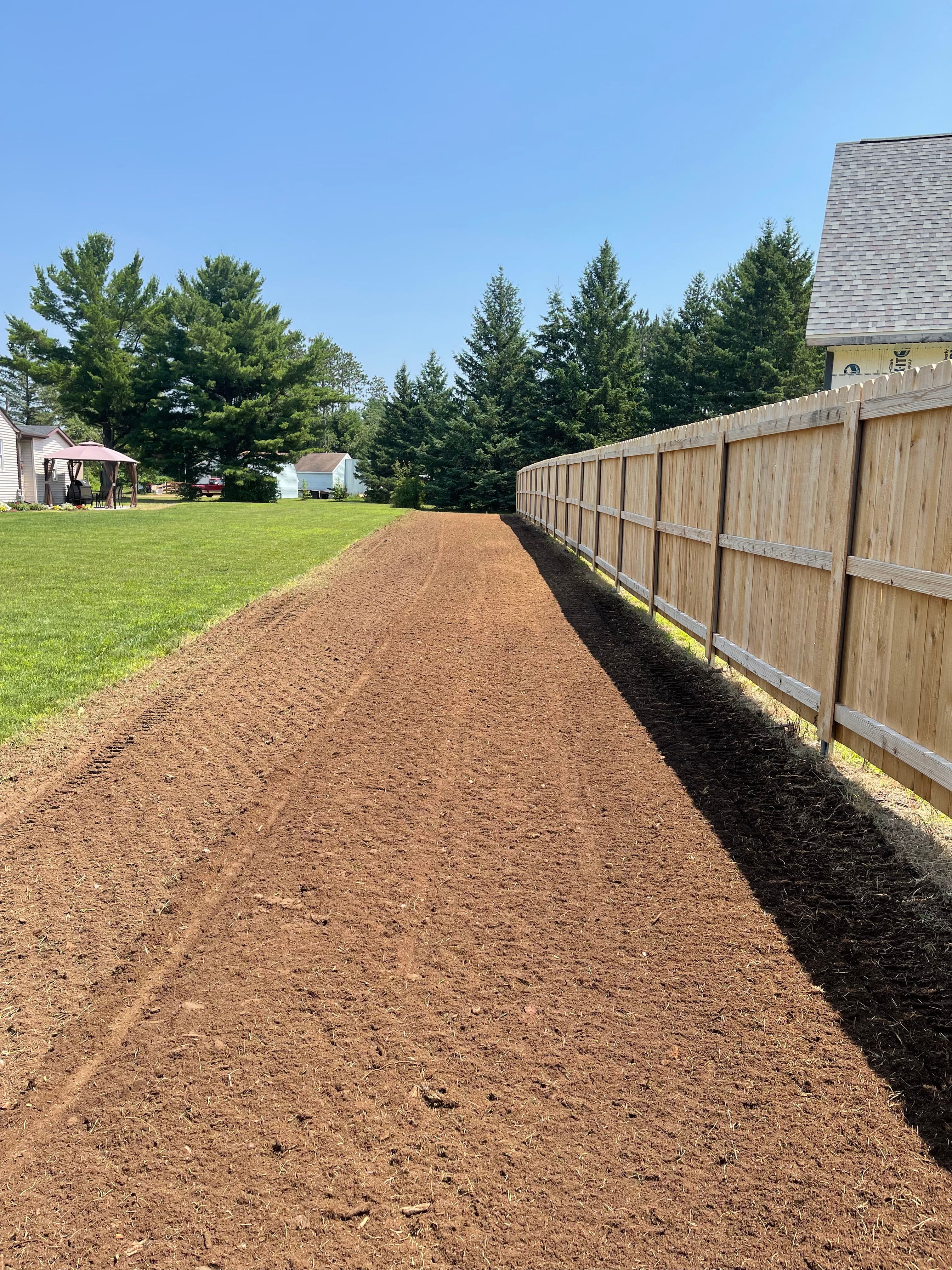 Dirt pathway alongside a wooden fence, bordered by trees and a green lawn under clear blue skies.